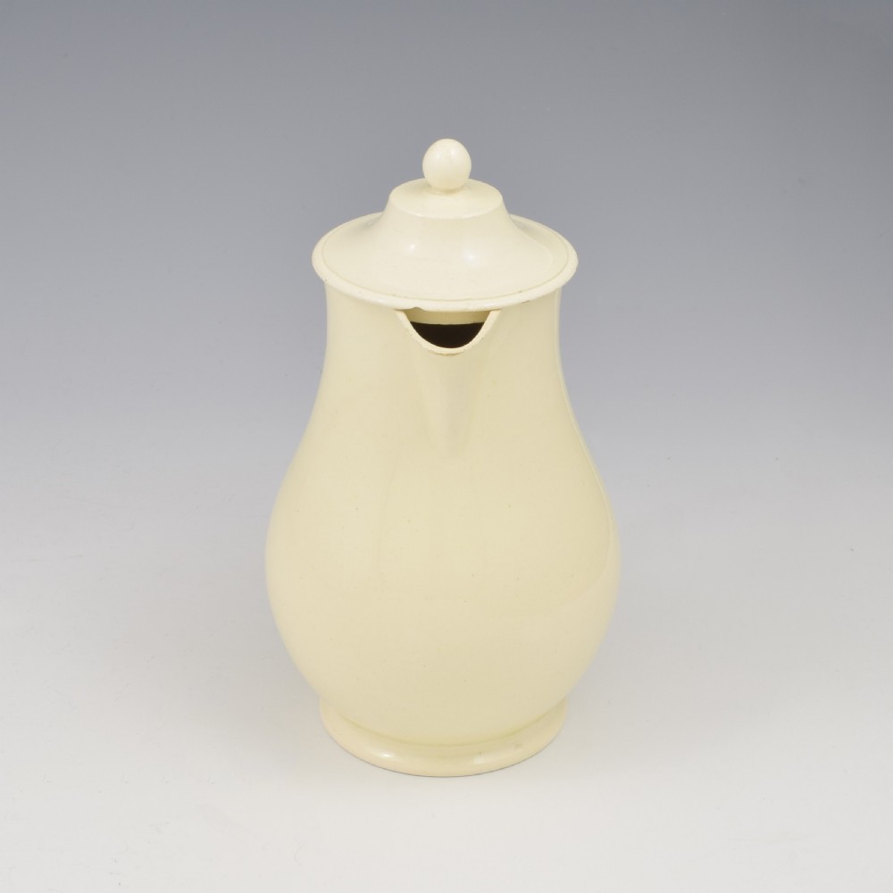 An 18th-century creamware hot water jug featuring a baluster-shaped earthenware body, a fitted dome cover with a ball finial, and a loop handle. View X