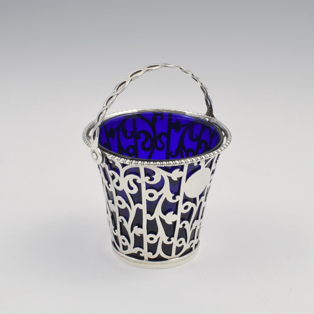 An 18th-century Georgian pierced sterling silver cream pail featuring intricate foliage scrollwork, a swing handle, and a removable cobalt blue glass liner. View II
