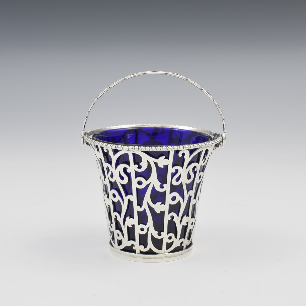 An 18th-century Georgian pierced sterling silver cream pail featuring intricate foliage scrollwork, a swing handle, and a removable cobalt blue glass liner. View V