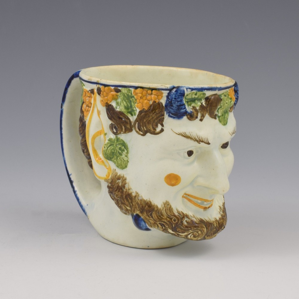 A late 18th-century pearlware ceramic mug formed as a satyrs head with relief-moulded beard, pointed ears, and grapevines in underglaze colours. View II