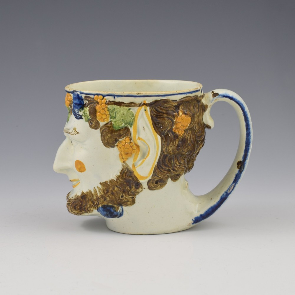 A late 18th-century pearlware ceramic mug formed as a satyrs head with relief-moulded beard, pointed ears, and grapevines in underglaze colours. View V