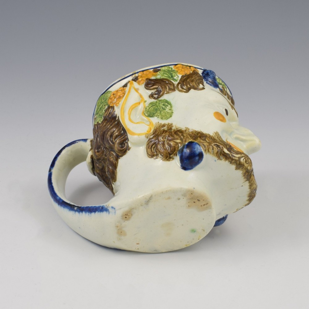 A late 18th-century pearlware ceramic mug formed as a satyrs head with relief-moulded beard, pointed ears, and grapevines in underglaze colours. View IX