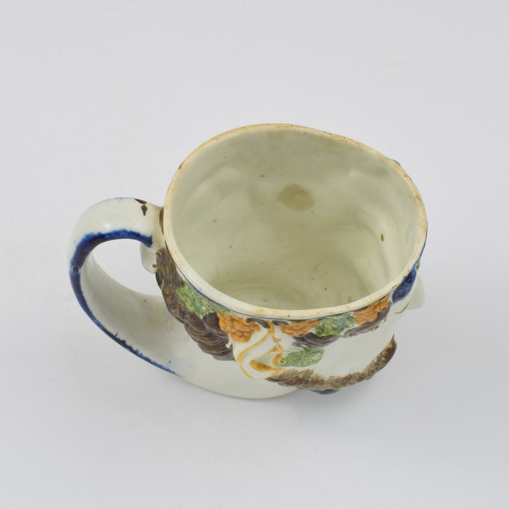 A late 18th-century pearlware ceramic mug formed as a satyrs head with relief-moulded beard, pointed ears, and grapevines in underglaze colours. View X