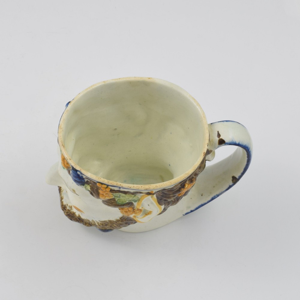 A late 18th-century pearlware ceramic mug formed as a satyrs head with relief-moulded beard, pointed ears, and grapevines in underglaze colours. View XI