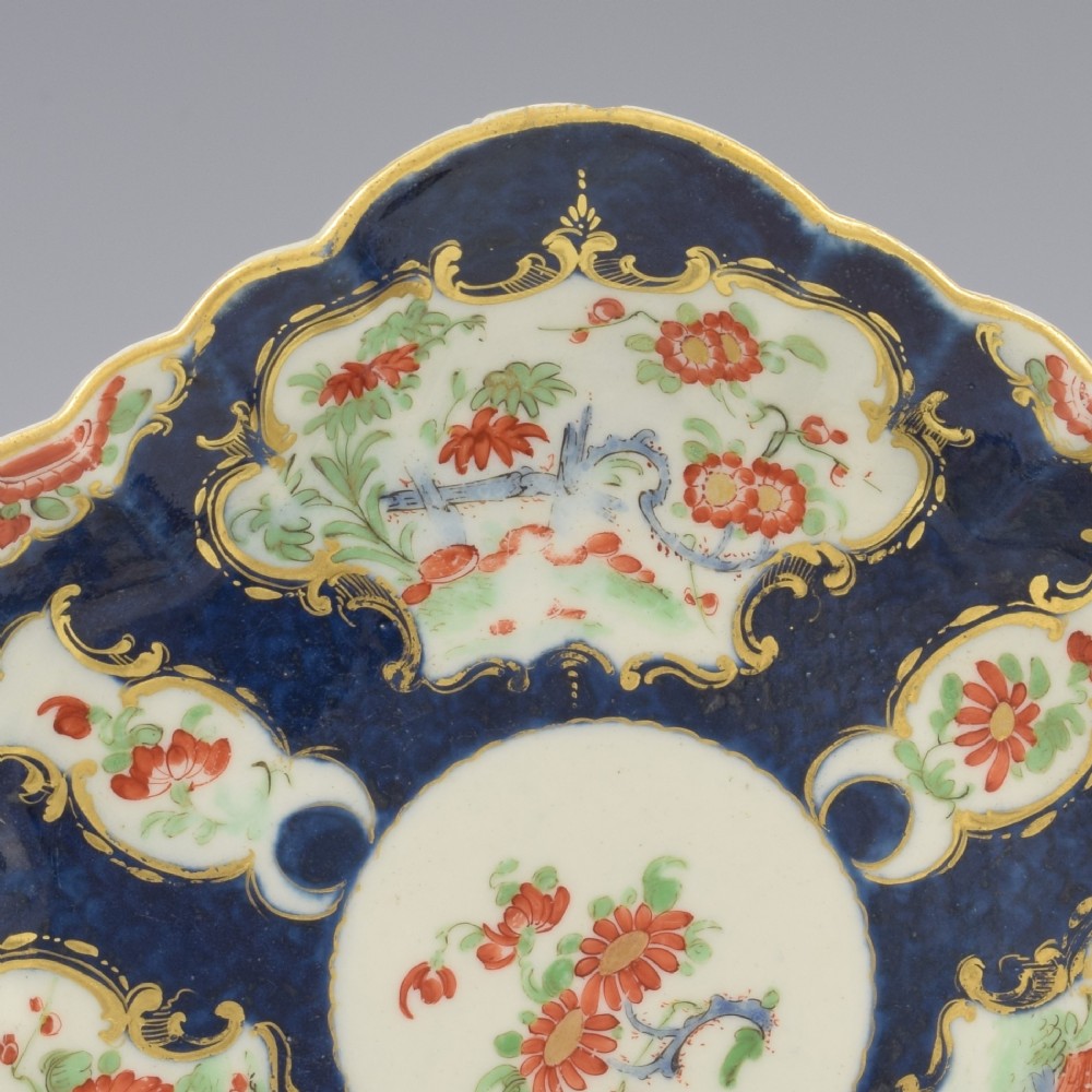 A scalloped Dr Wall Worcester porcelain teapot stand featuring a hand-painted blue scale ground, gilded rococo cartouches, and colourful Kakiemon-style floral motifs. View II