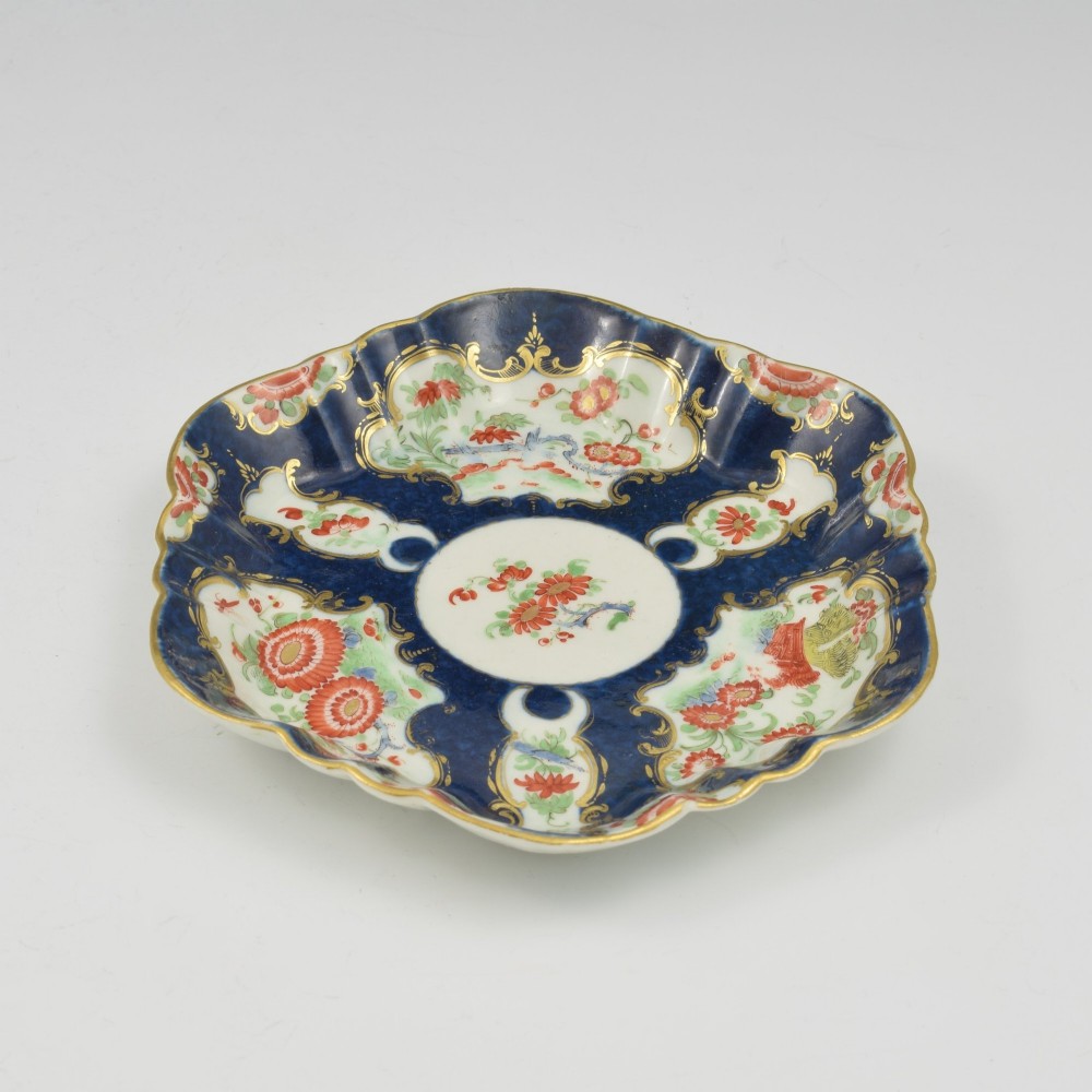 A scalloped Dr Wall Worcester porcelain teapot stand featuring a hand-painted blue scale ground, gilded rococo cartouches, and colourful Kakiemon-style floral motifs. View VI