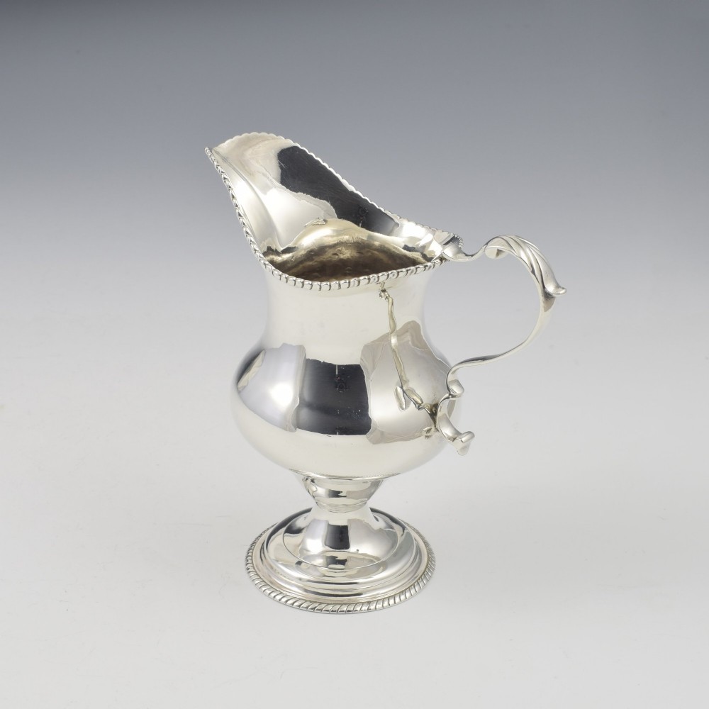 A Georgian sterling silver milk jug featuring a baluster body, pedestal foot, beaded rim, and double-scroll handle. View X