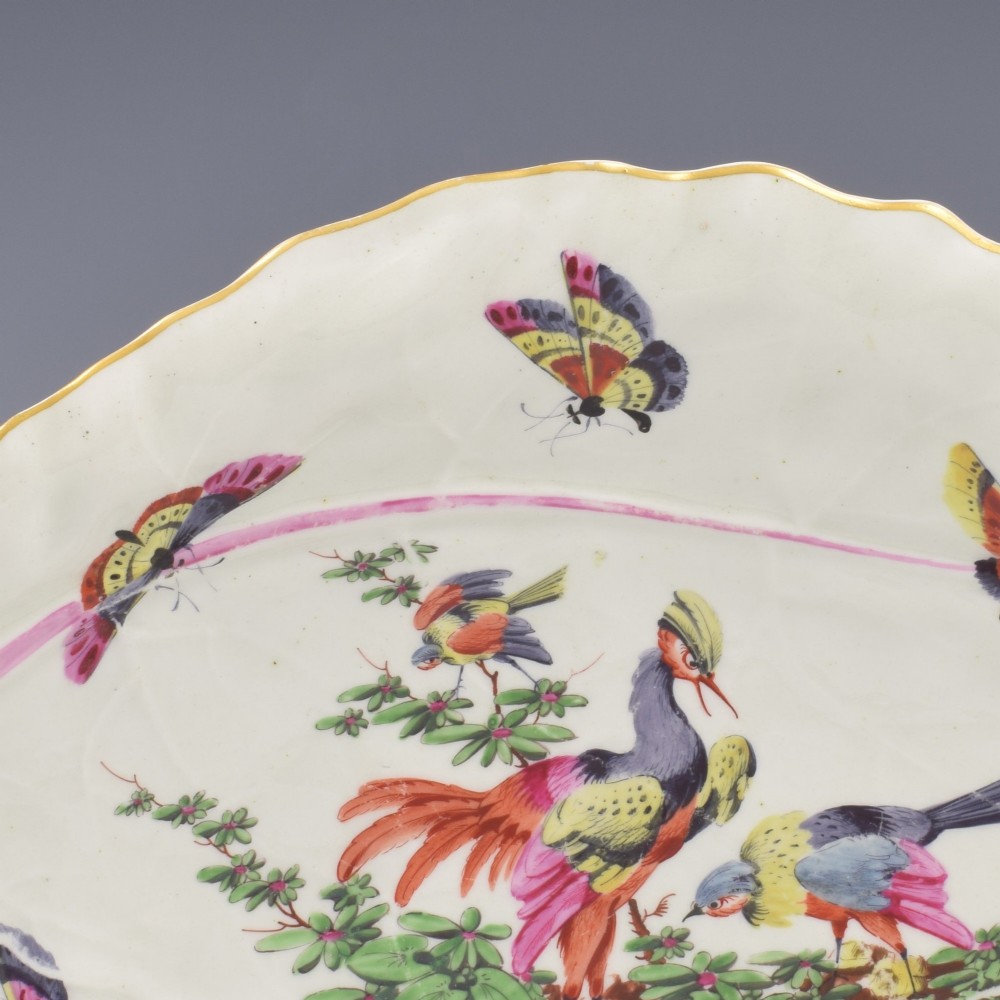 An eighteenth-century leaf-moulded soft-paste porcelain dish featuring central hand-painted fancy birds, surrounding butterflies, a pink stalk handle, and a gilded edge. View V
