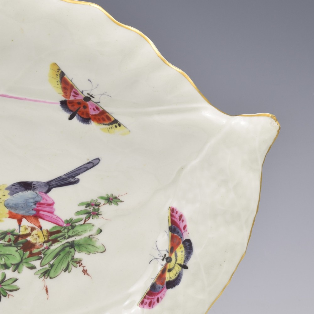 An eighteenth-century leaf-moulded soft-paste porcelain dish featuring central hand-painted fancy birds, surrounding butterflies, a pink stalk handle, and a gilded edge. View VI