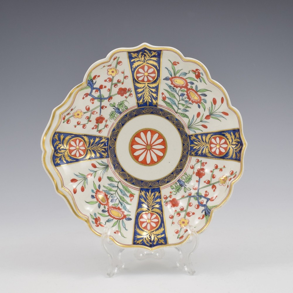 An 18th-century circular soft-paste porcelain dish with a scalloped rim, decorated in the Rich Queens pattern featuring cobalt blue gilt panels alternating with painted red and green floral motifs. View II
