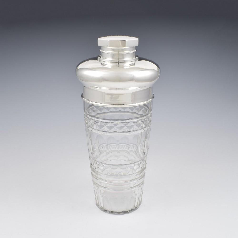 An Art Deco cocktail shaker featuring a clear cut glass body with geometric facet patterns and a polished sterling silver mount and octagonal cap.