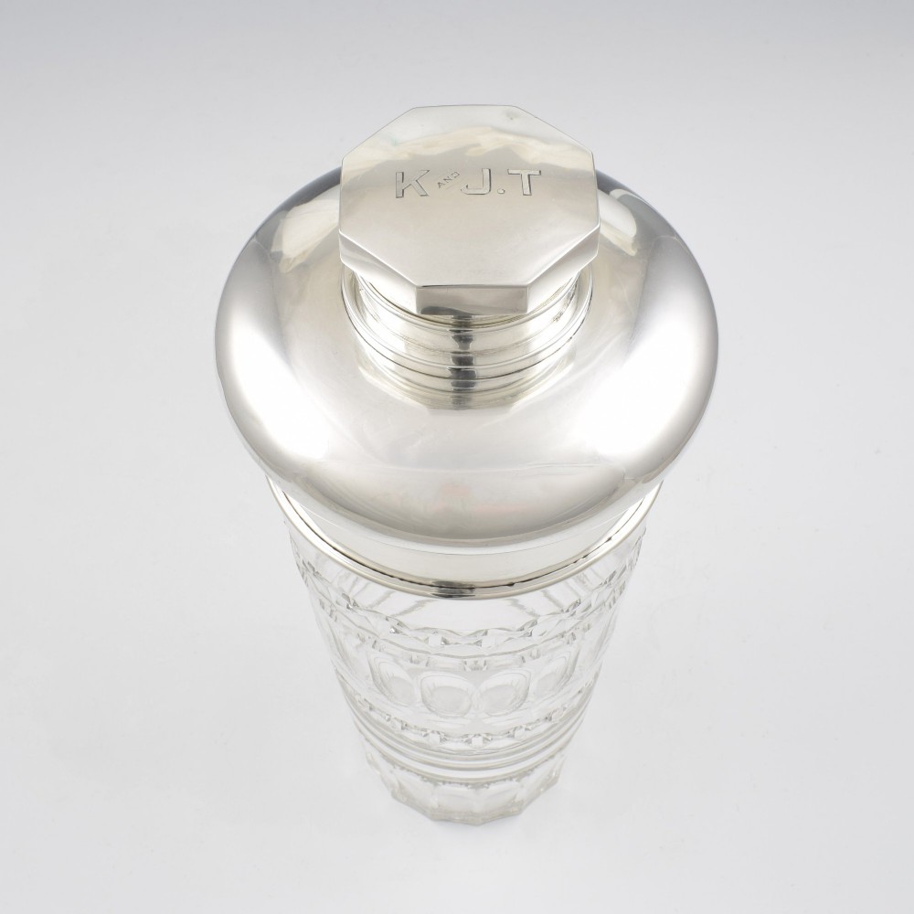 An Art Deco cocktail shaker featuring a clear cut glass body with geometric facet patterns and a polished sterling silver mount and octagonal cap. View V