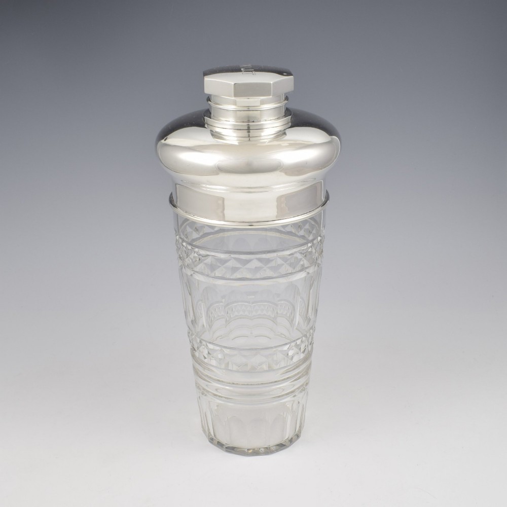 An Art Deco cocktail shaker featuring a clear cut glass body with geometric facet patterns and a polished sterling silver mount and octagonal cap. View X