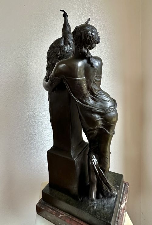 fig 1. A 19th-century bronze sculpture depicting a draped female figure holding a small mirror up to a peacock perched on a pillar, mounted on a red marble base.