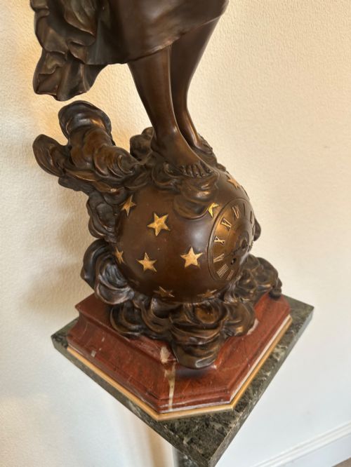fig 1. A dark bronze sculpture depicting a draped female figure standing atop a starry globe and mounted on a dark red marble base.