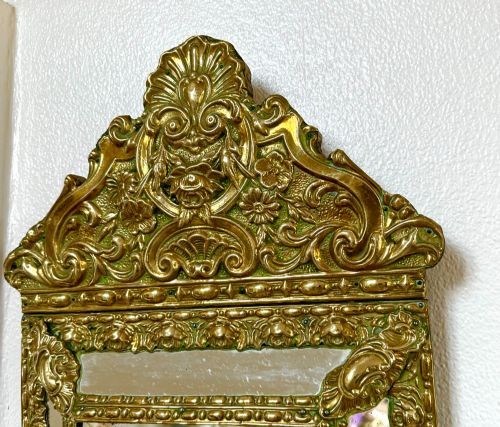fig 1. An antique repousse brass marginal mirror featuring an ornate floral pediment crest and bevelled glass borders.