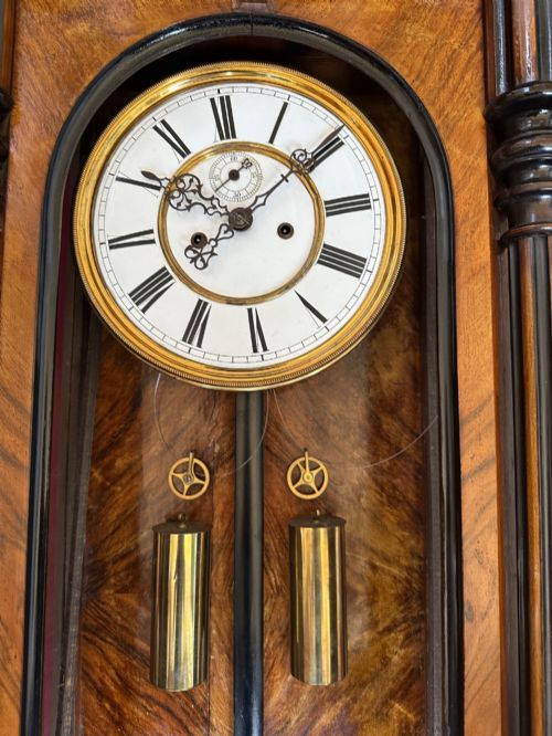 fig 1. Late 19th-century Victorian walnut wall clock with ebonised columns, top cresting with a carved head motif, two brass weights, pendulum, and a white enamelled Roman numeral dial.