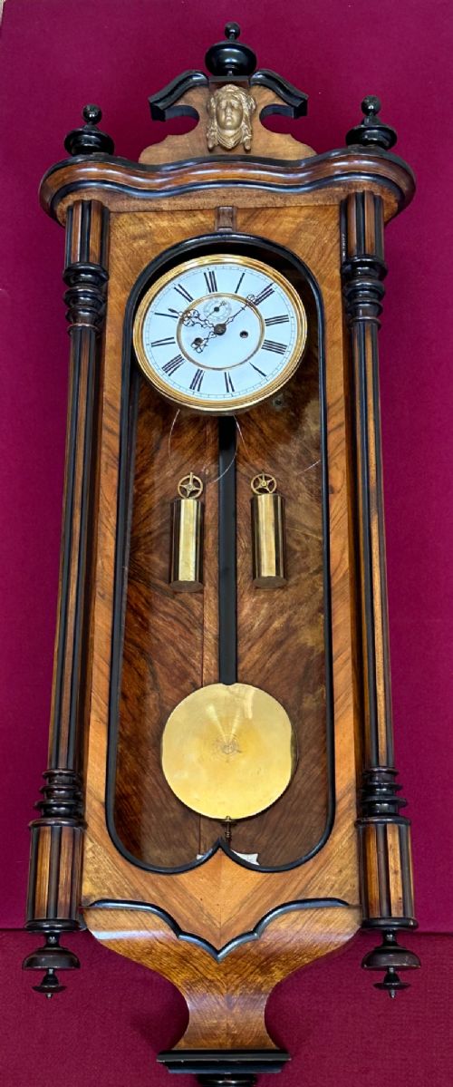 Late 19th-century Victorian walnut wall clock with ebonised columns, top cresting with a carved head motif, two brass weights, pendulum, and a white enamelled Roman numeral dial. View VI