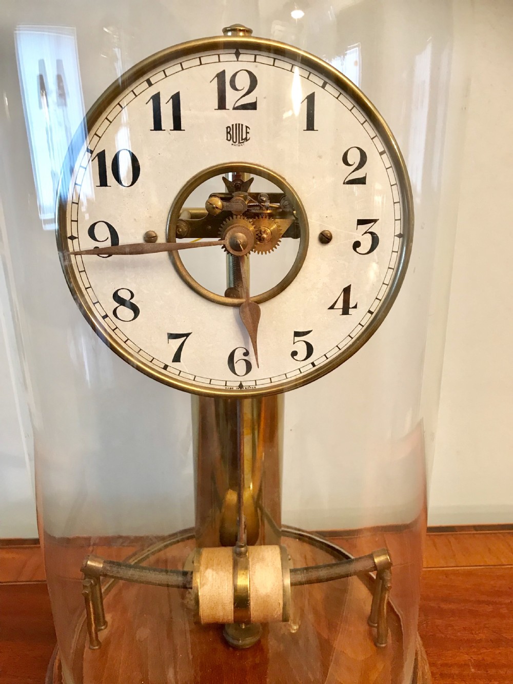An antique Bulle electric clock displaying an open brass movement with a white dial, enclosed beneath a tall clear glass dome on a wooden base. View IV