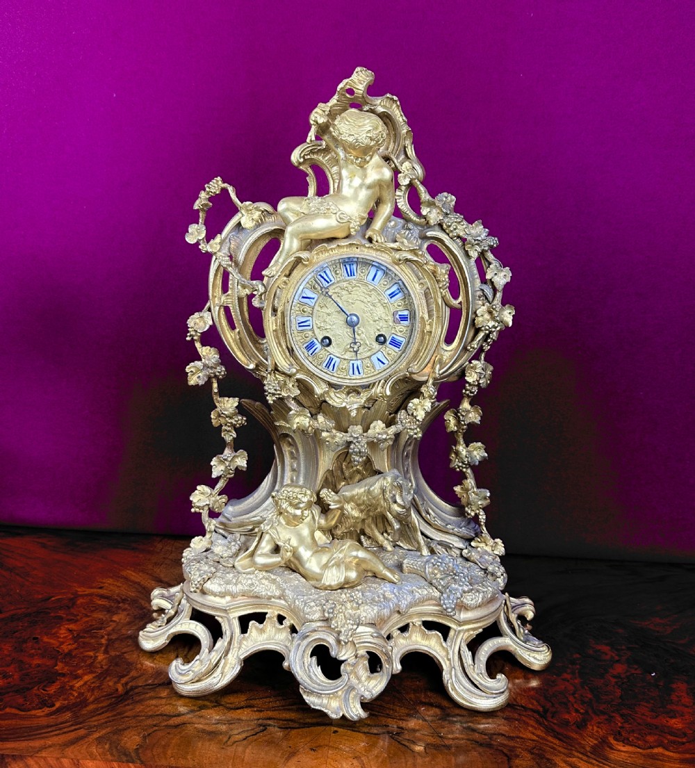 An ornate 19th-century French gilt bronze mantel clock featuring sculptural cherubs, grapevine garlands, and an enamel Roman numeral dial face.