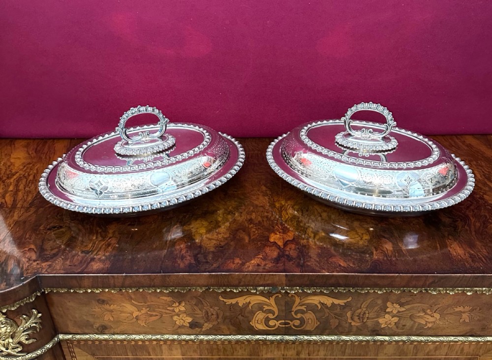 A pair of oval silver-plated entree dishes with beaded rims, engraved lids, and detachable handles, displayed on a burr wood surface.