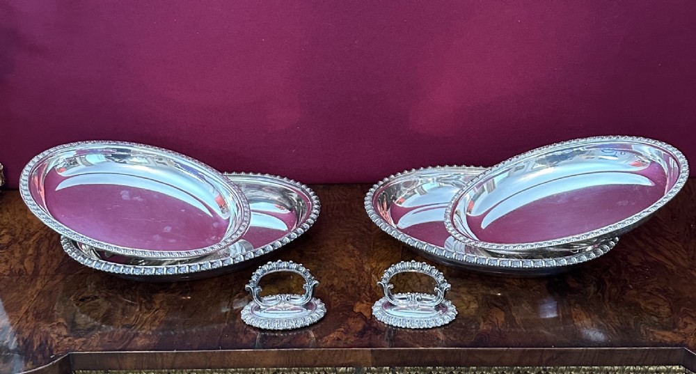 A pair of oval silver-plated entree dishes with beaded rims, engraved lids, and detachable handles, displayed on a burr wood surface. View V
