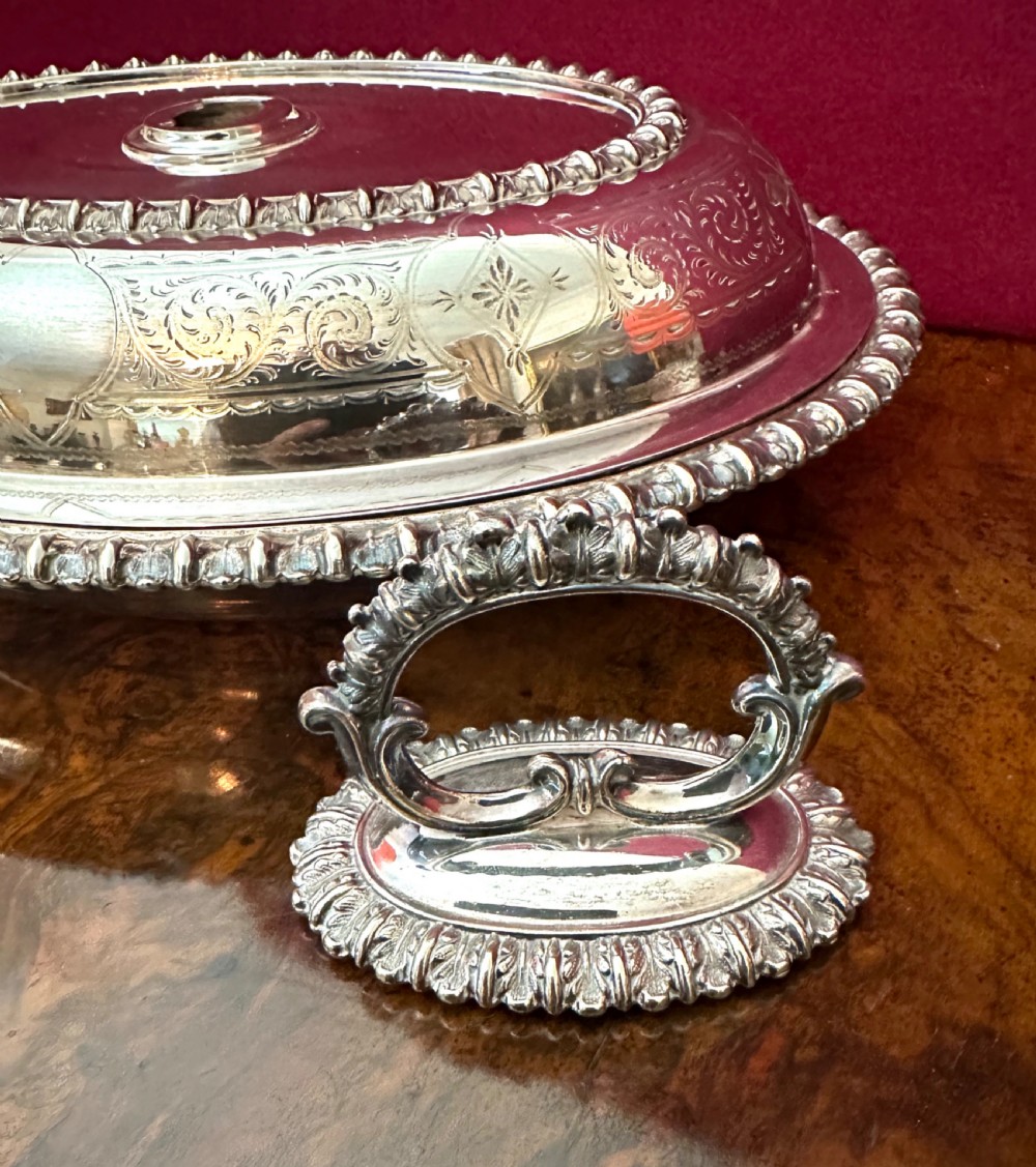 A pair of oval silver-plated entree dishes with beaded rims, engraved lids, and detachable handles, displayed on a burr wood surface. View VI