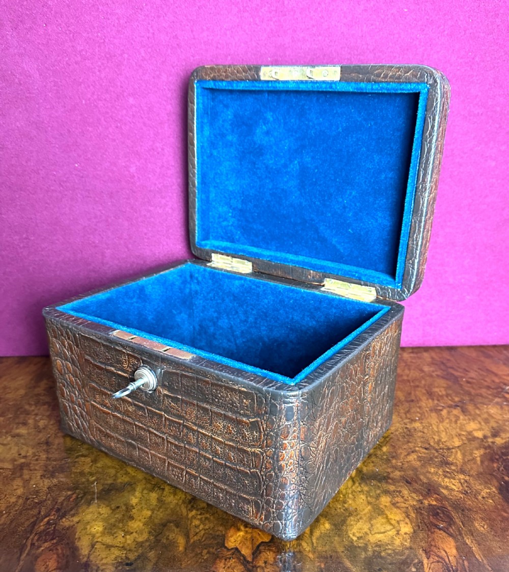 A rectangular dark brown crocodile leather jewellery box shown open, displaying a bright blue velvet interior lining and a metal key inserted into the front lock.