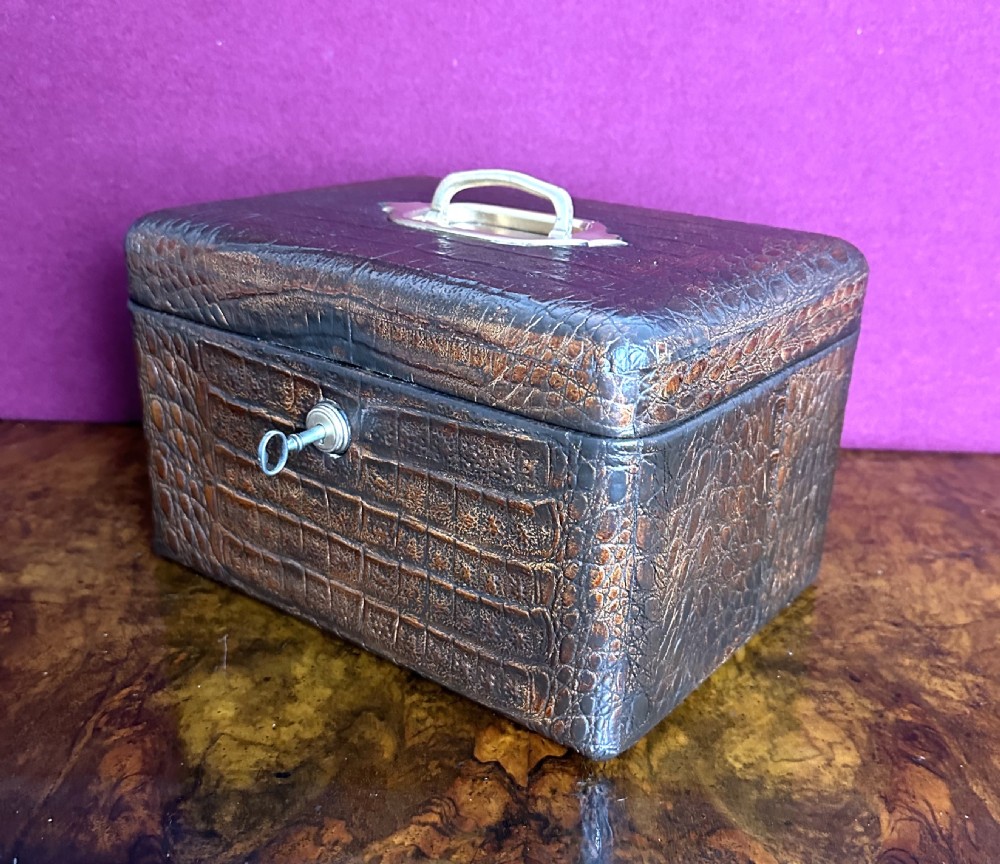 A rectangular dark brown crocodile leather jewellery box shown open, displaying a bright blue velvet interior lining and a metal key inserted into the front lock. View V