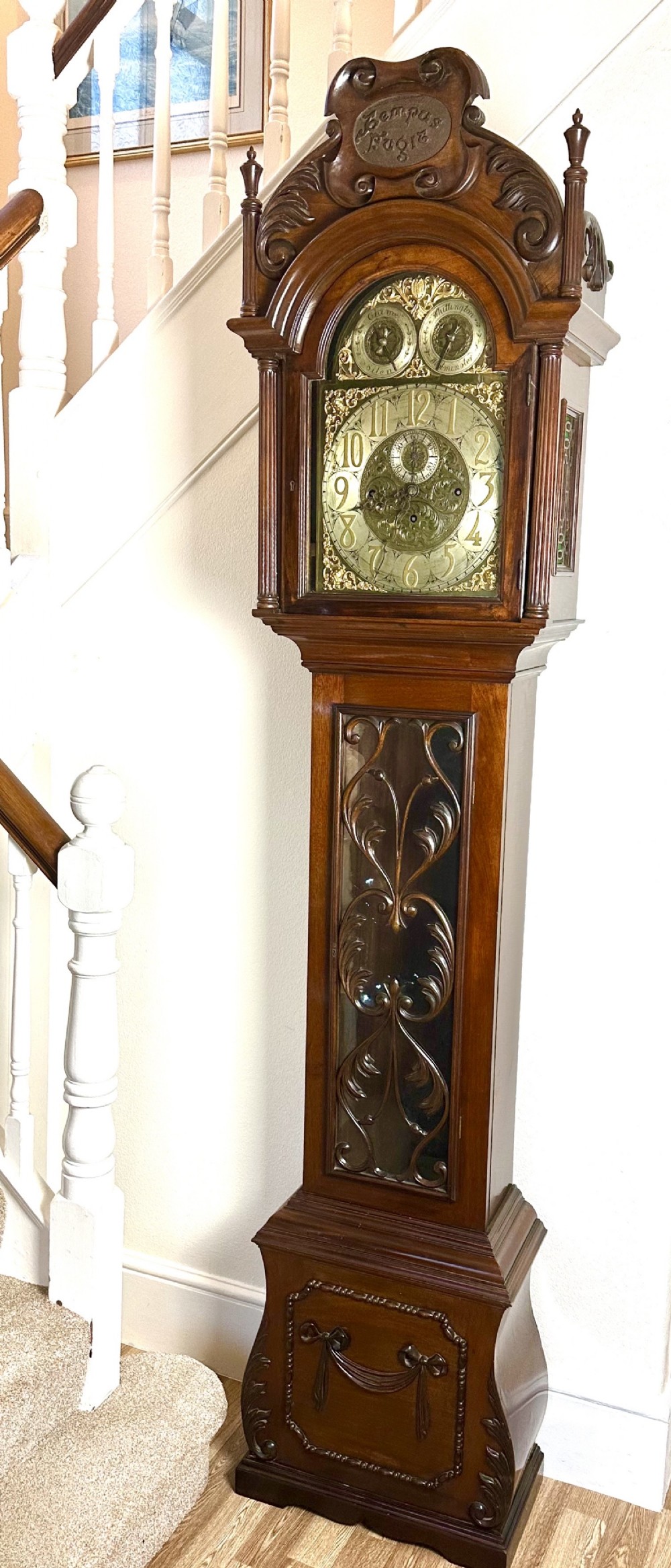 A carved mahogany longcase clock featuring an ornate brass dial, scrolled pediment with a Tempus Fugit plaque, and decorative wood tracery glass panel.
