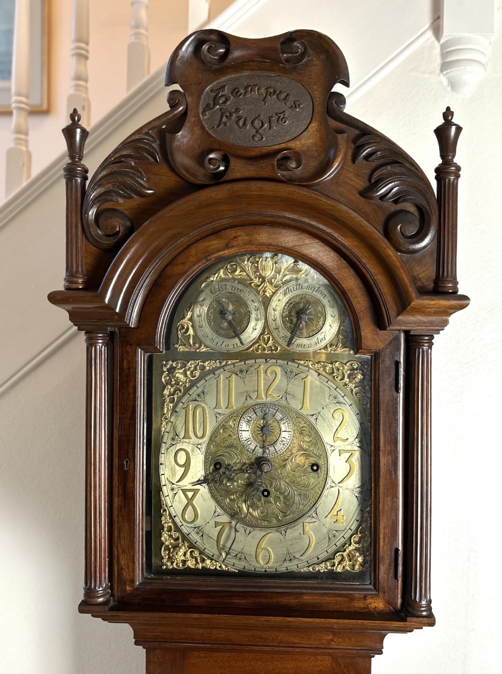 A carved mahogany longcase clock featuring an ornate brass dial, scrolled pediment with a Tempus Fugit plaque, and decorative wood tracery glass panel. View III