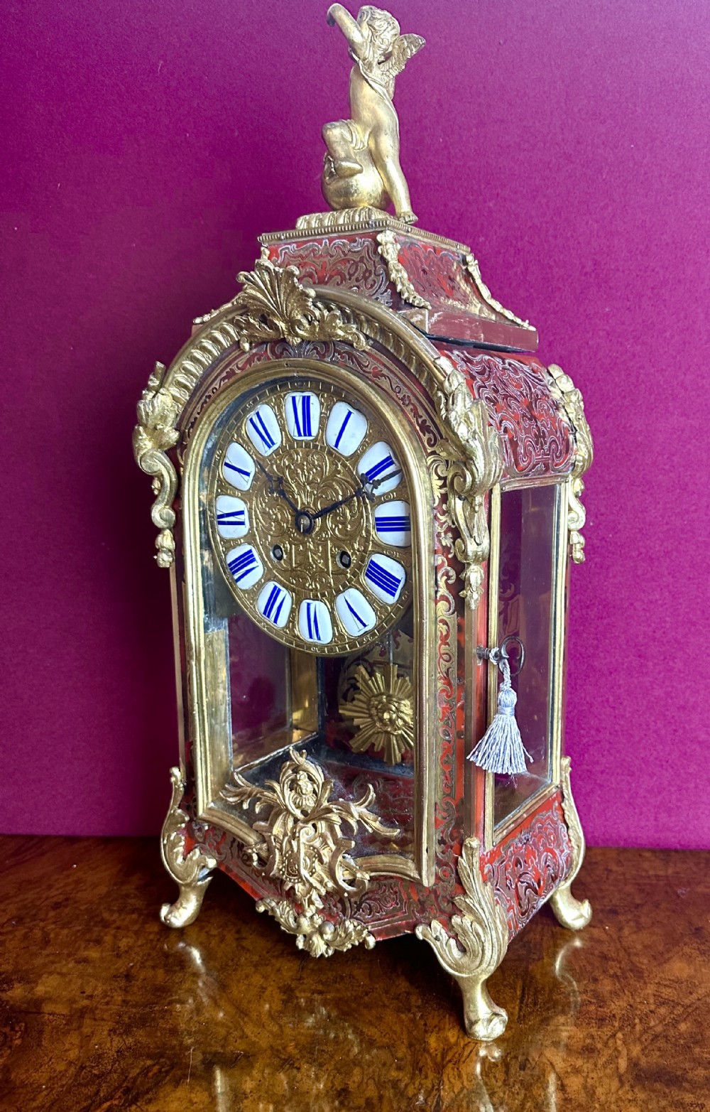An 19th-century French Boulle mantel clock featuring red tortoiseshell with brass marquetry, gilt ormolu mounts, an enamel cartouche dial, and a winged cherub finial.