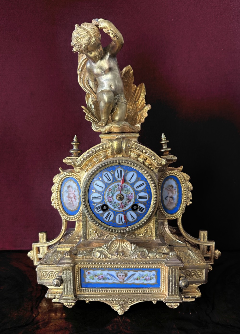 A French Rococo Revival gilt bronze mantel clock surmounted by a sculpted cherub, featuring blue porcelain panels and an enamel Roman numeral dial.