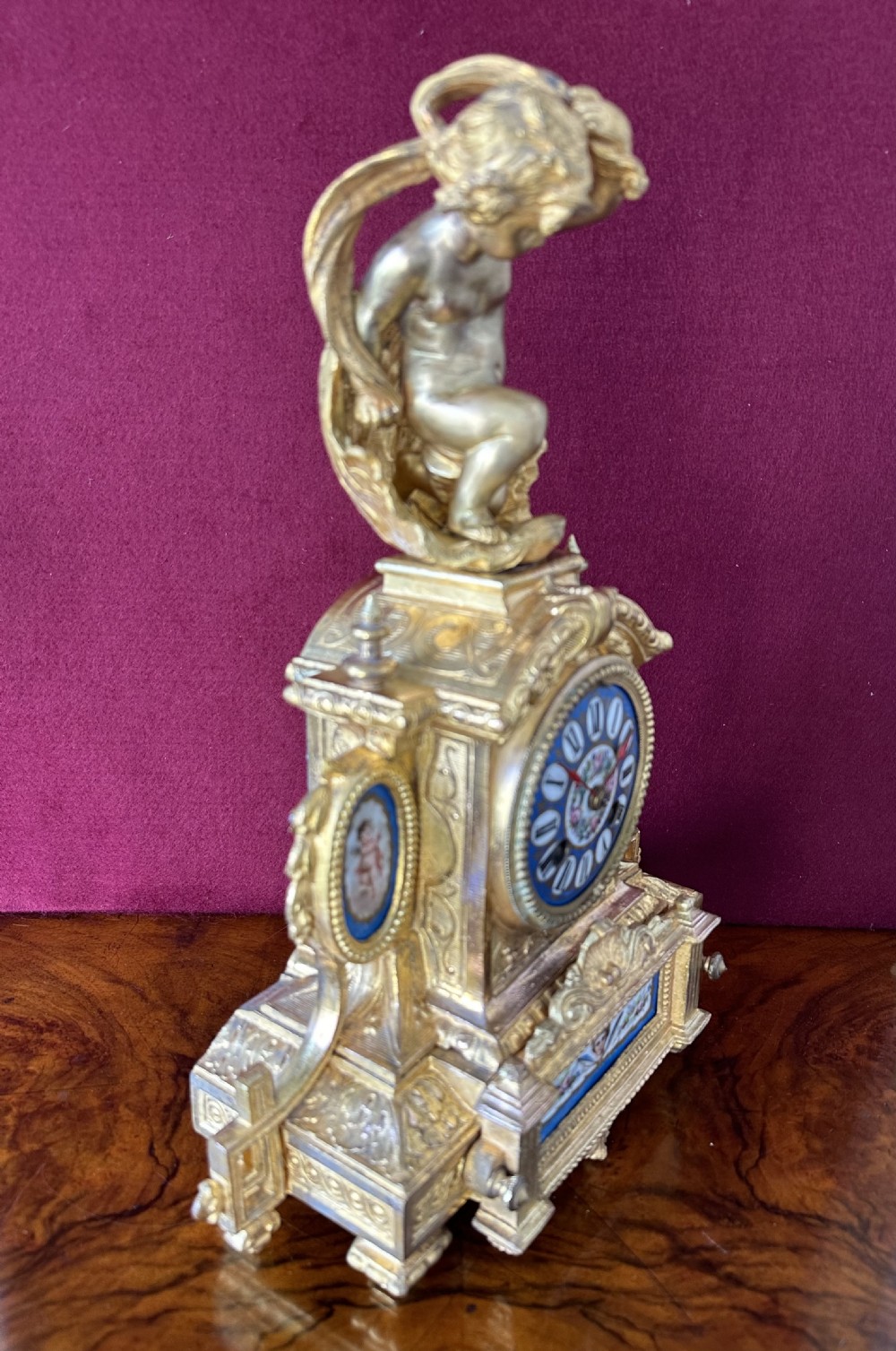 A French Rococo Revival gilt bronze mantel clock surmounted by a sculpted cherub, featuring blue porcelain panels and an enamel Roman numeral dial. View VI