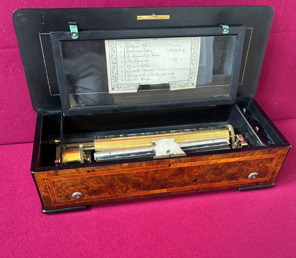 An open Victorian music box featuring a burr walnut inlaid wooden case, an interior glass cover displaying a brass cylinder mechanism, and an original paper tune sheet inside the lid.