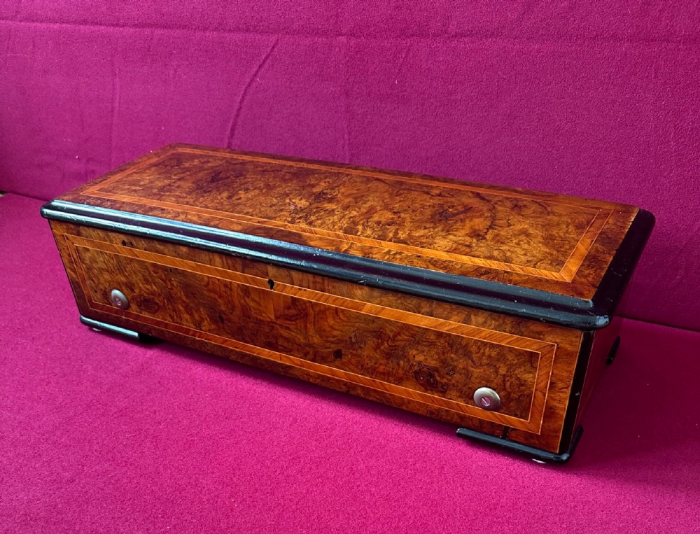 An open Victorian music box featuring a burr walnut inlaid wooden case, an interior glass cover displaying a brass cylinder mechanism, and an original paper tune sheet inside the lid. View IV