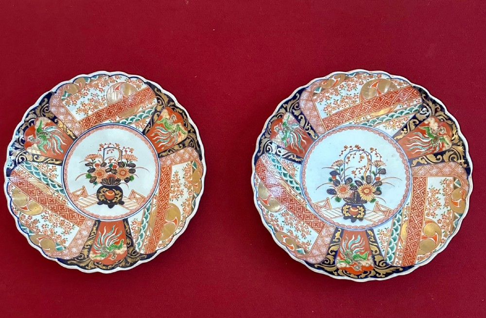 A pair of circular Japanese Imari porcelain chargers featuring scalloped edges, cobalt blue and iron-red painted panels, and central flower basket motifs with gilded accents.