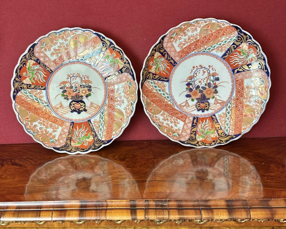 A pair of circular Japanese Imari porcelain chargers featuring scalloped edges, cobalt blue and iron-red painted panels, and central flower basket motifs with gilded accents. View V