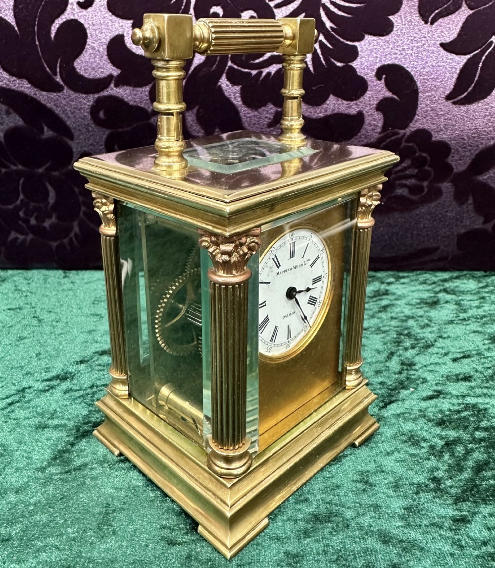 An antique brass carriage clock featuring Corinthian column corners, a top carrying handle, glass side panels, and a white enamel dial with Roman numerals. View II