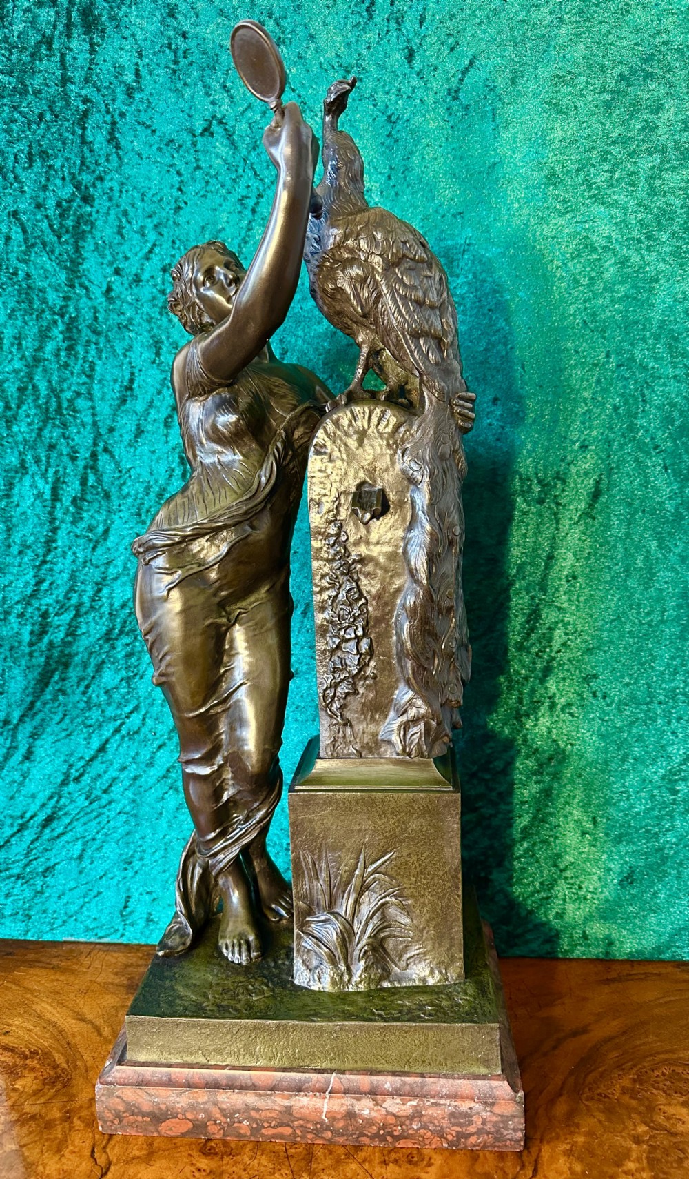 A 19th-century bronze sculpture depicting a draped female figure holding a small mirror up to a peacock perched on a pillar, mounted on a red marble base.