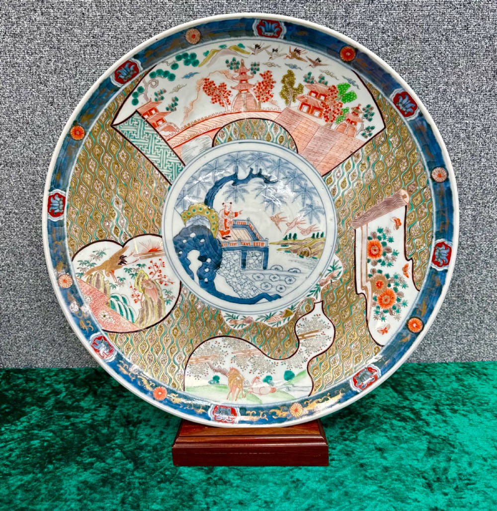 A large circular Japanese Imari porcelain charger decorated with underglaze cobalt blue and overglaze terracotta red enamels depicting landscape scenes and geometric borders.