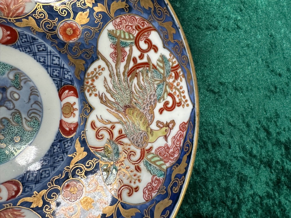 A circular Japanese Imari porcelain plate decorated in cobalt blue, iron-red, and gold accents around panels featuring phoenixes and blossom trees. View III