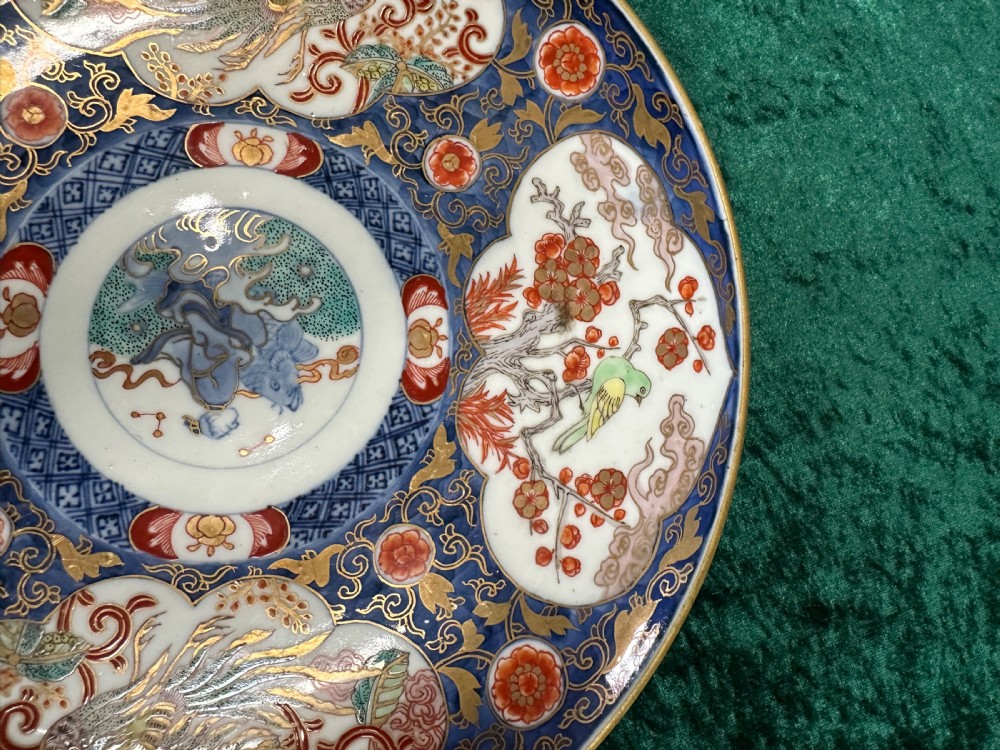 A circular Japanese Imari porcelain plate decorated in cobalt blue, iron-red, and gold accents around panels featuring phoenixes and blossom trees. View V