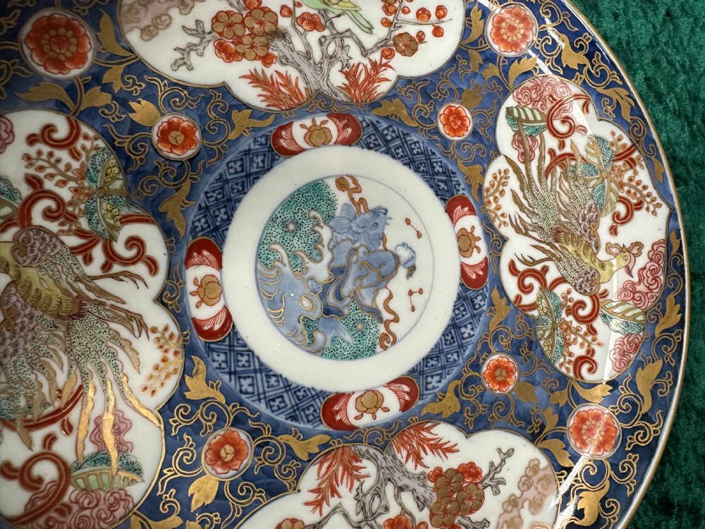 A circular Japanese Imari porcelain plate decorated in cobalt blue, iron-red, and gold accents around panels featuring phoenixes and blossom trees. View VI