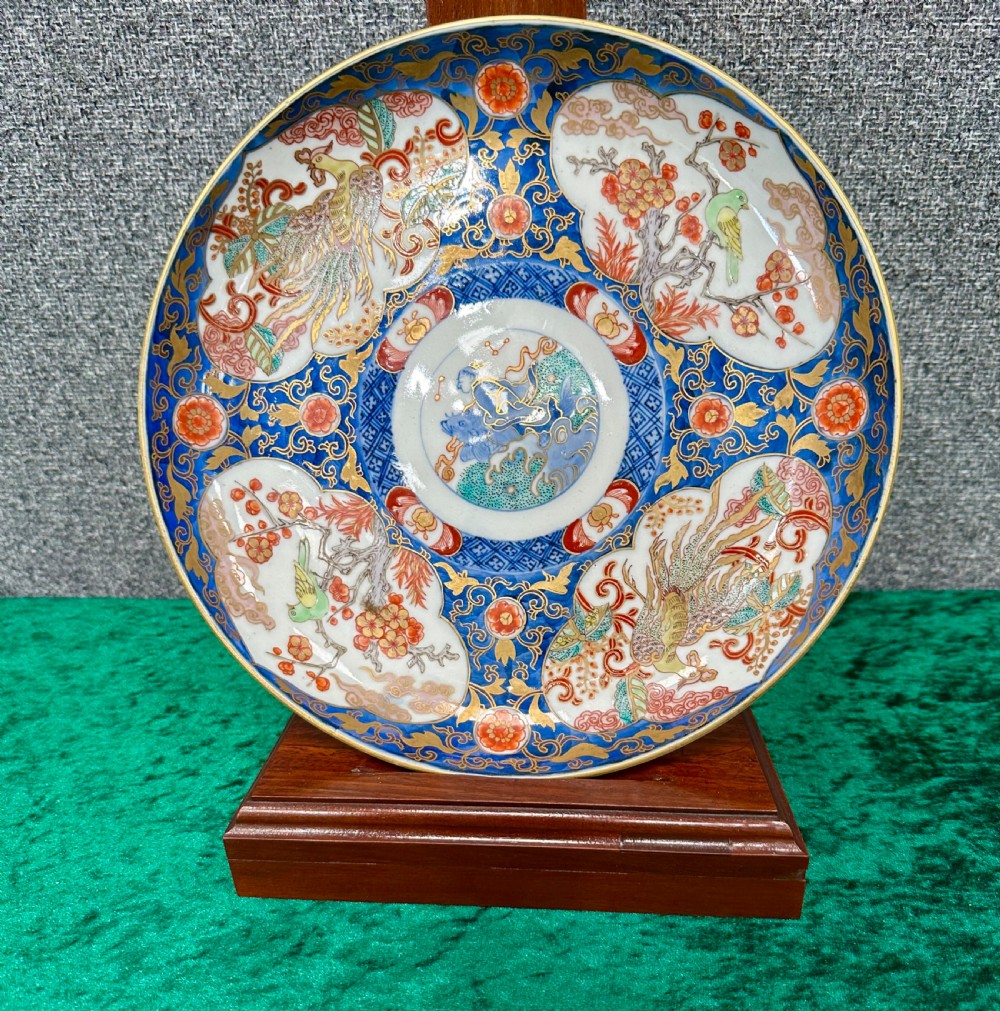 A circular Japanese Imari porcelain plate decorated in cobalt blue, iron-red, and gold accents around panels featuring phoenixes and blossom trees. View IX