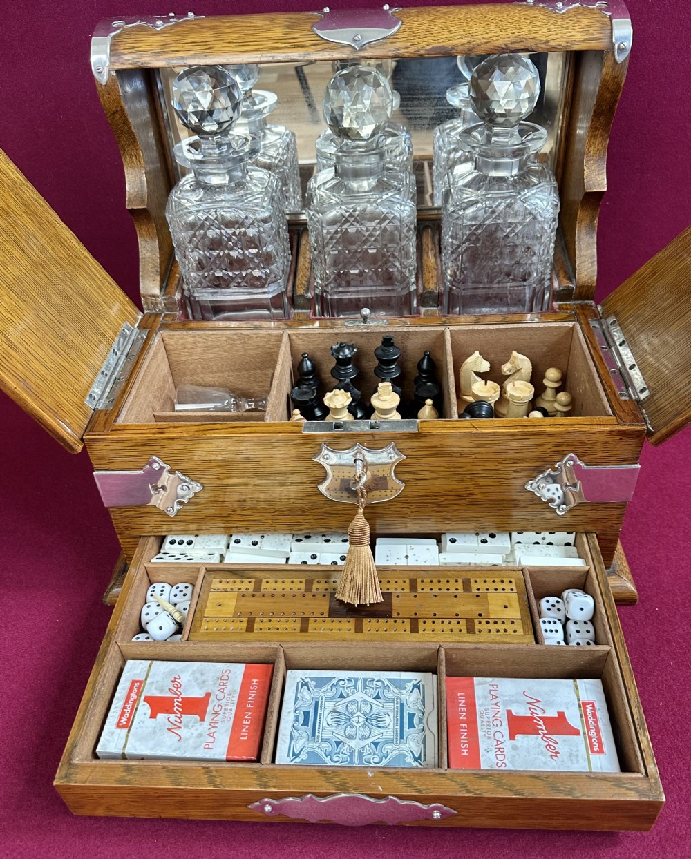 An open Edwardian oak games compendium displaying three cut crystal decanters, chess pieces, cribbage board, dominoes, and playing cards.
