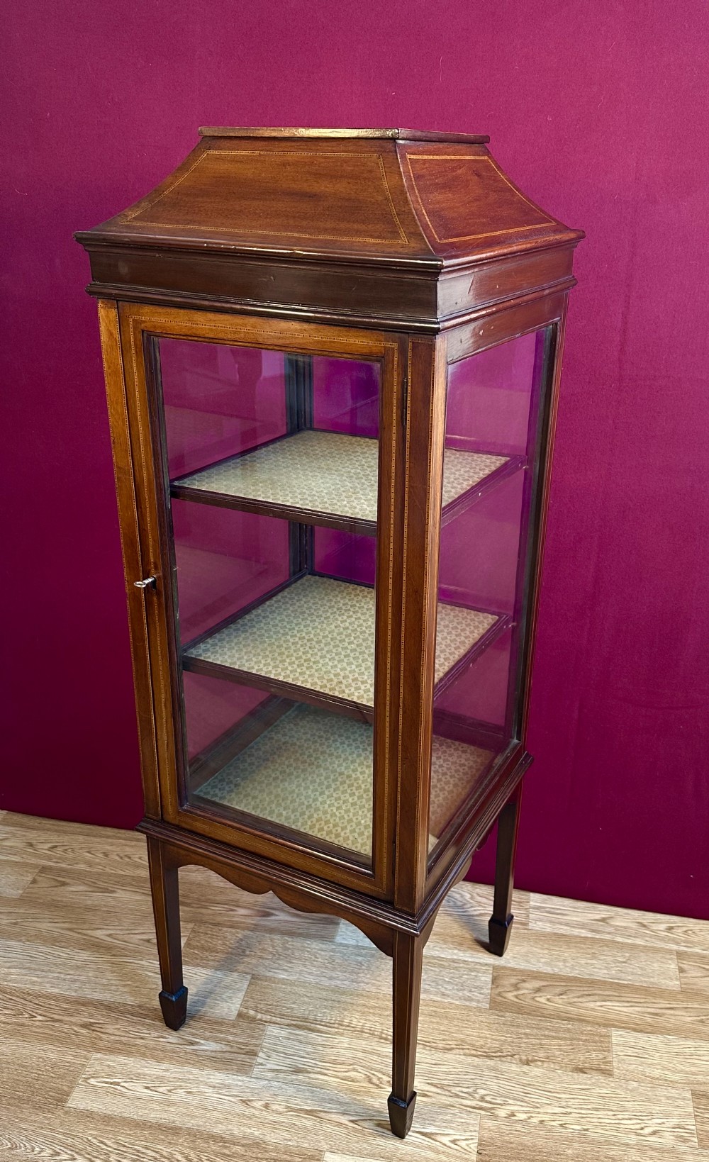An Edwardian inlaid mahogany pedestal vitrine cabinet featuring glass panels on four sides, three fabric-lined shelves, a single glazed door, and a caddy top roof design.