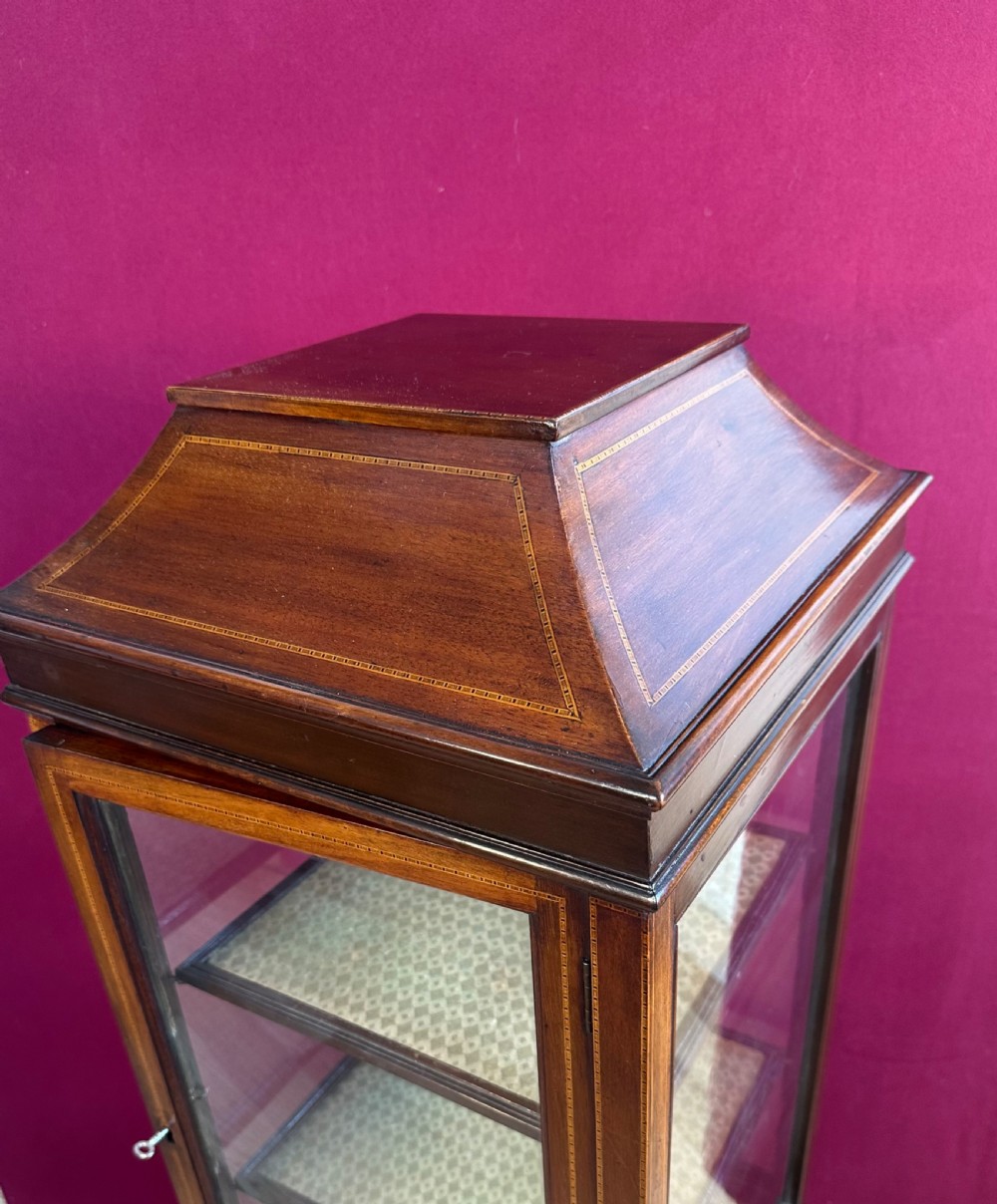An Edwardian inlaid mahogany pedestal vitrine cabinet featuring glass panels on four sides, three fabric-lined shelves, a single glazed door, and a caddy top roof design. View VIII