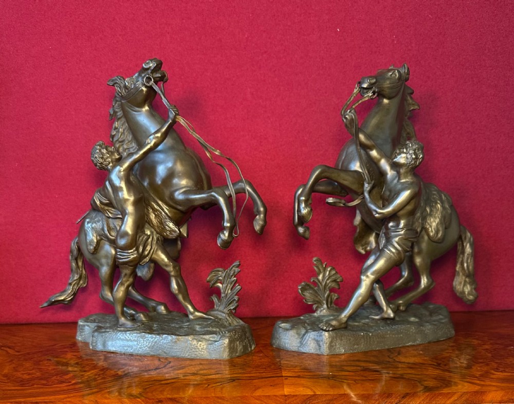 A pair of patinated bronze sculptures depicting rearing horses restrained by standing groom figures on naturalistic bases.