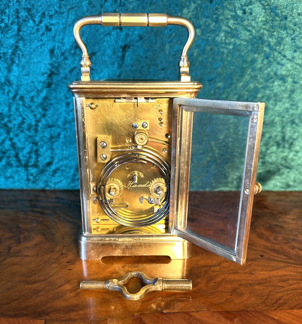 A rectangular brass carriage clock featuring glass side panels, a white enamel dial with black Roman numerals, and a top carrying handle. View III